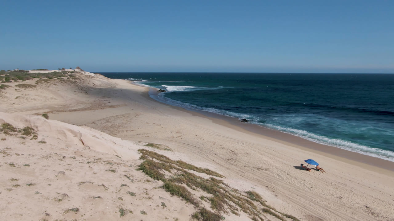Wide aerial beach with sand dunes, blue ocean, surf, and a blue umbrella.