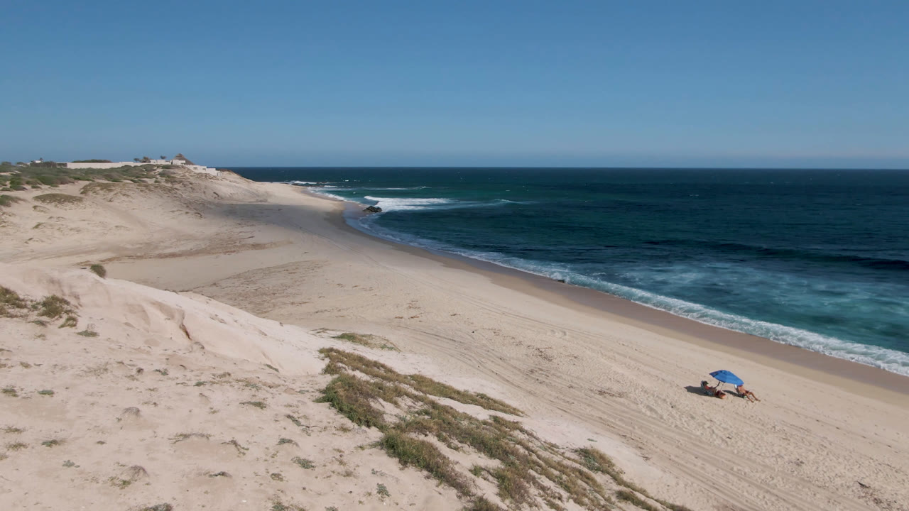 High wide view of Shipwrecks Beach in Los Cabos with the blue umbrella near the lower sand, dunes and sparse vegetation at left, and a broad band of teal water and breaking surf at right.