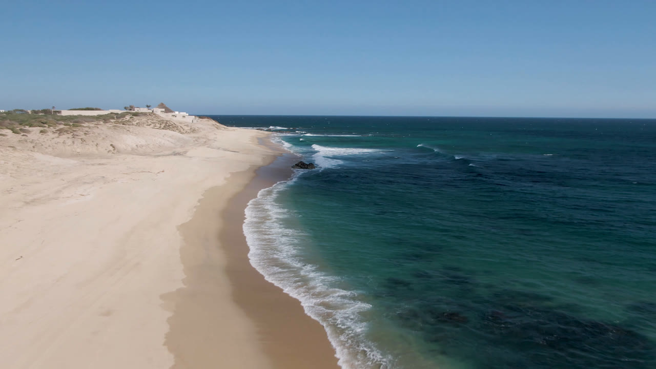 High oblique view at Shipwrecks Beach Los Cabos, Baja California Sur, with the shoreline shifting lower in frame, foamy waves crossing the sand, dark water patches offshore, and no visible people.