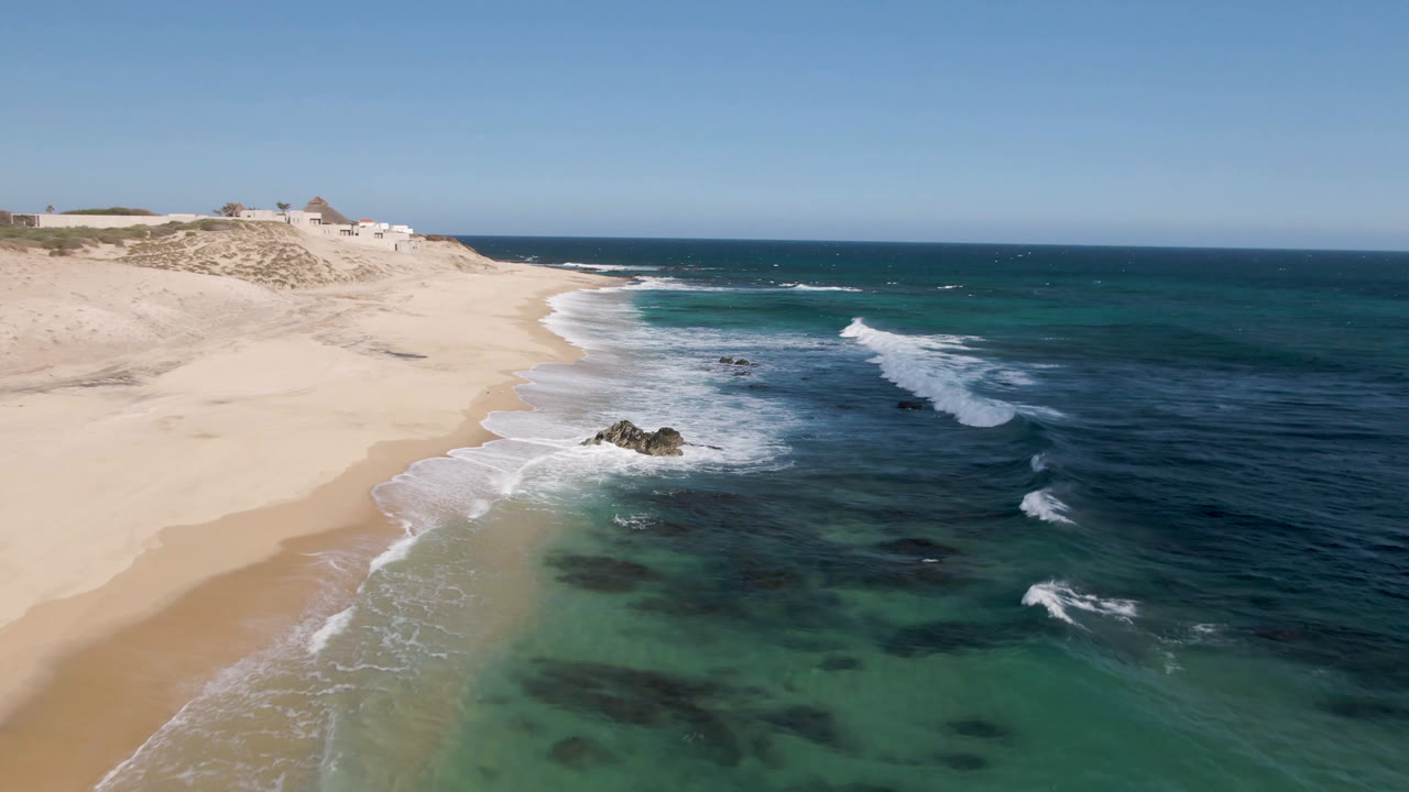 Drone view at Shipwrecks Beach with white surf pushing toward a broad sandy shoreline, rocks near the center foreground, dunes behind the beach, and no visible people.