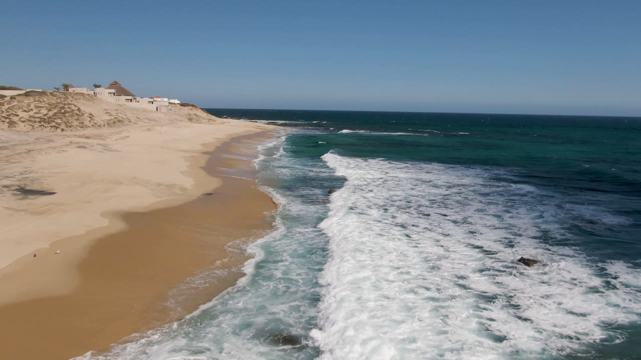 Aerial frame over Shipwrecks Beach in Los Cabos with tan sand curving left, white surf entering from the lower right, distant dunes and small structures, clear sky, and no visible people.