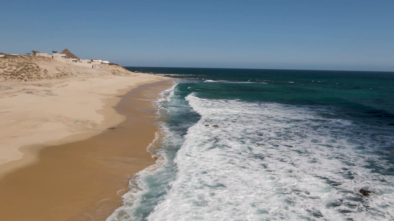 Aerial view at Shipwrecks Beach in Los Cabos with a broad sheet of whitewater pushing toward the sand, darker ocean beyond, small rocks in the surf, distant dune structures, and no visible people.