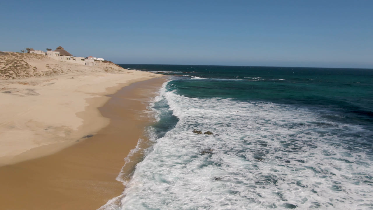 Aerial coastline view with surf, rocks, sand, and open blue water.