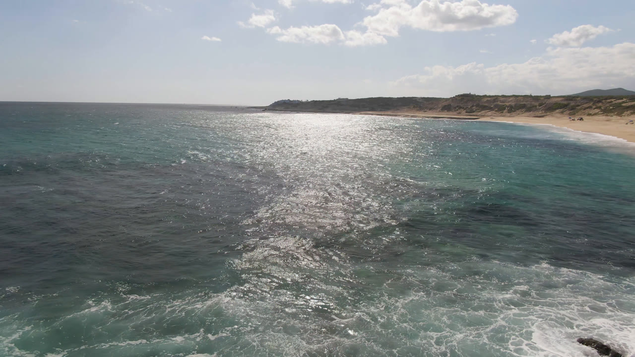 Aerial view over sunlit ocean toward a sandy beach and low dunes