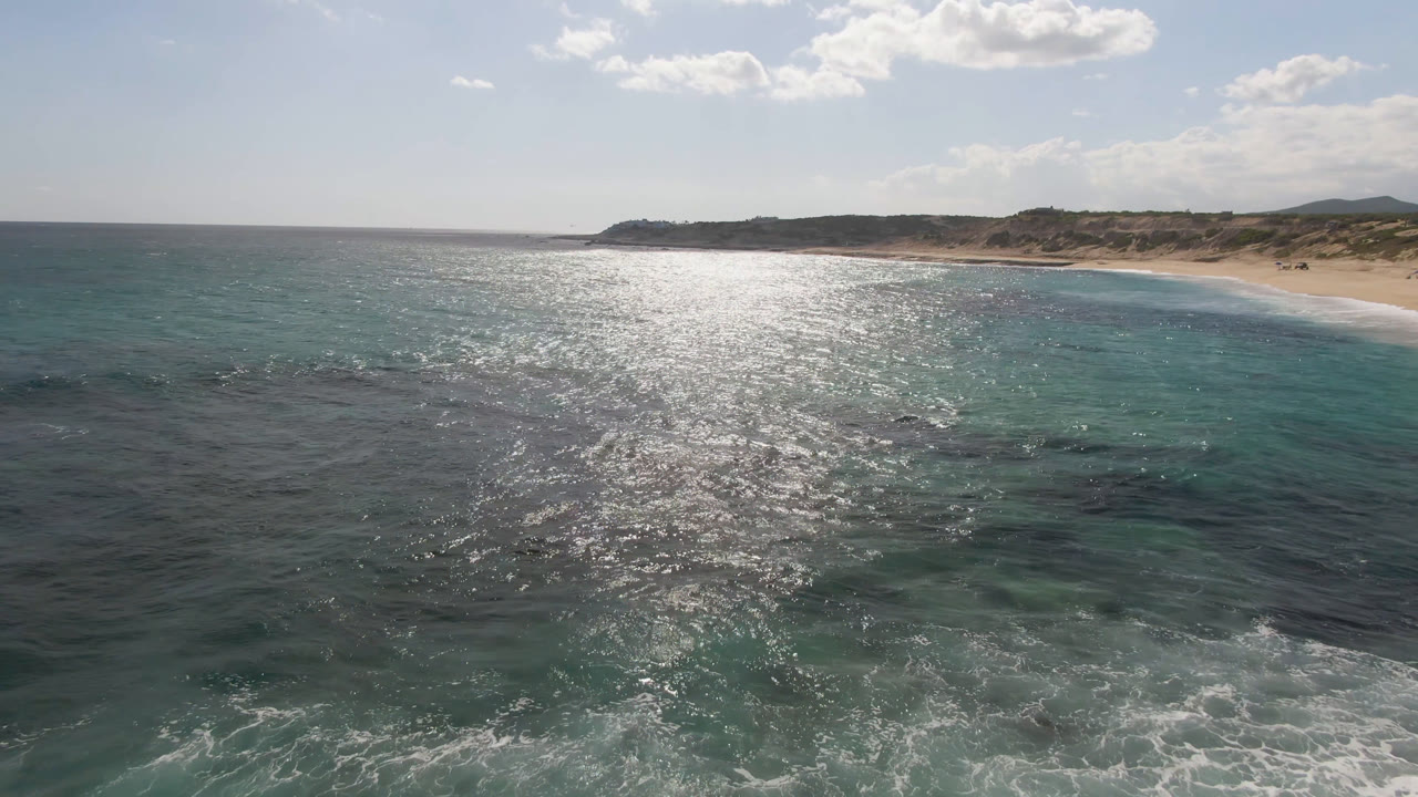At 0:01, the Shipwrecks Beach Los Cabos aerial keeps the shimmering ocean centered, with darker reef-like patches below the surface, pale sand beyond, and no visible people.