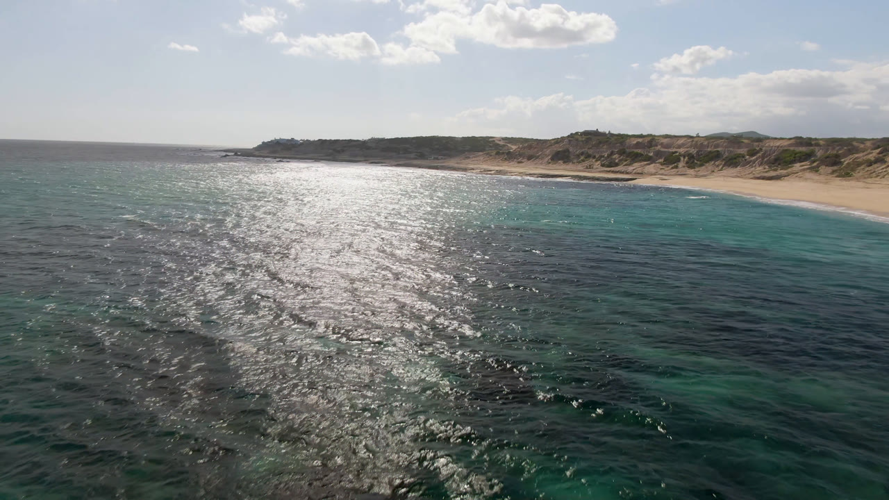 Aerial frame over Shipwrecks Beach in Los Cabos, with bright sun reflection across dark and turquoise water, sandy shoreline beyond, low dunes under scattered clouds, and no visible people.