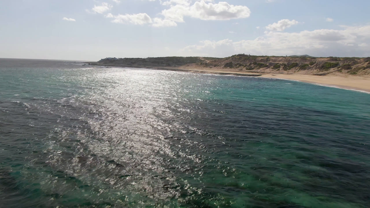 Aerial view at Shipwrecks Beach Los Cabos showing a wide band of sun glare on rippled ocean, green-blue shallows near the sand, low scrub-covered dunes, scattered clouds, and no visible people.