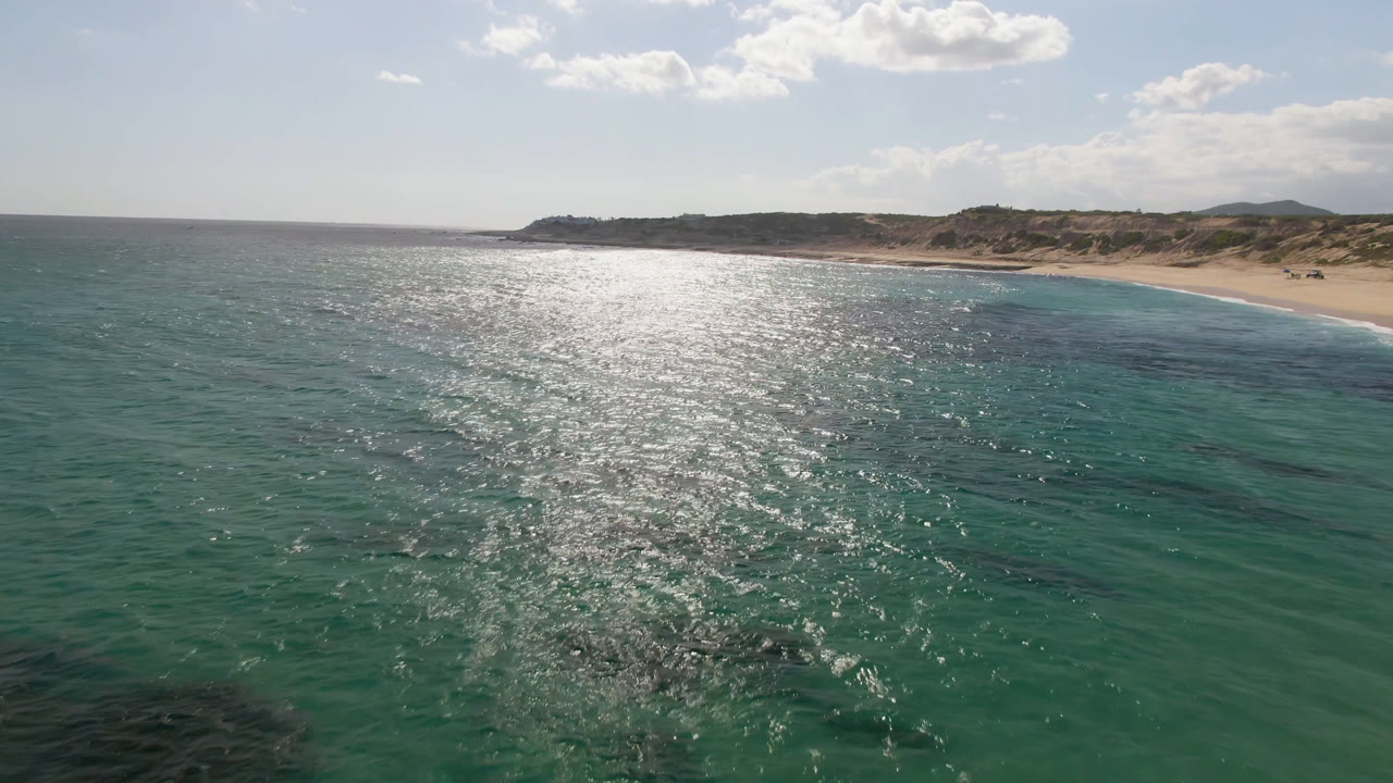 Aerial frame over Shipwrecks Beach Los Cabos, with sun glare across green-blue water, a sandy shoreline in the distance, low desert hills behind it, and no visible people.