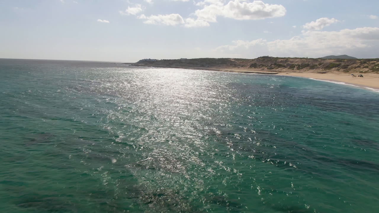 Aerial view at Shipwrecks Beach Los Cabos, showing a broad band of sunlit ocean, darker underwater patches, sandy beach on the right, desert hills beyond, and no visible people.