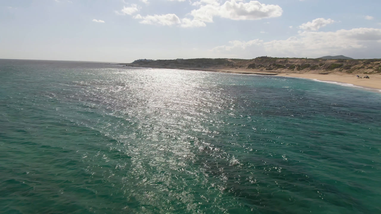 Drone view of ocean surface with a curving beach and desert terrain beyond.