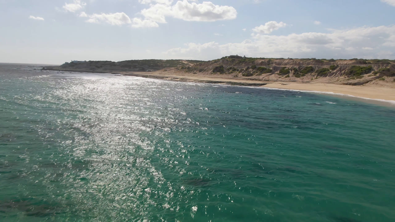 Opening aerial frame over turquoise water at Shipwrecks Beach in Los Cabos, with sun glitter on the left side, sandy shore and low bluffs beyond, and no visible people.