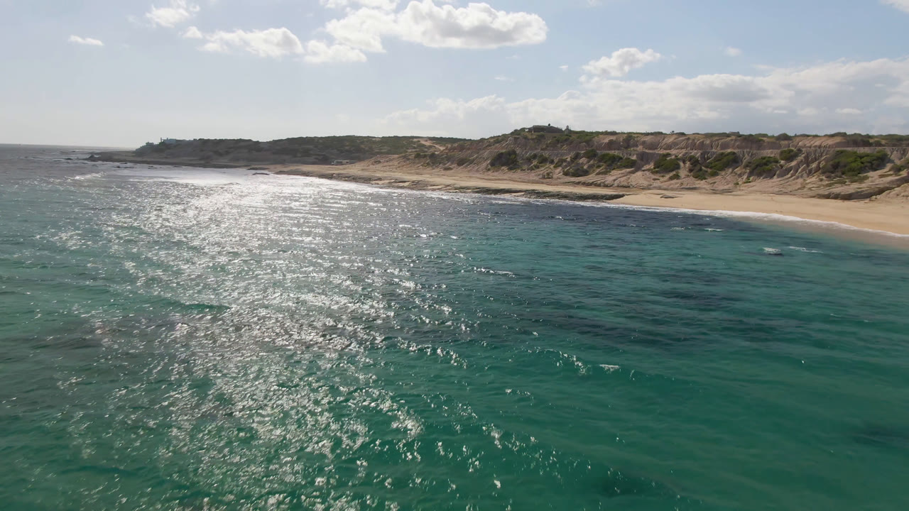 Aerial view at Shipwrecks Beach in Los Cabos where bright reflection spreads across the water, the sandy beach curves behind it, and no visible people appear on shore or in the sea.