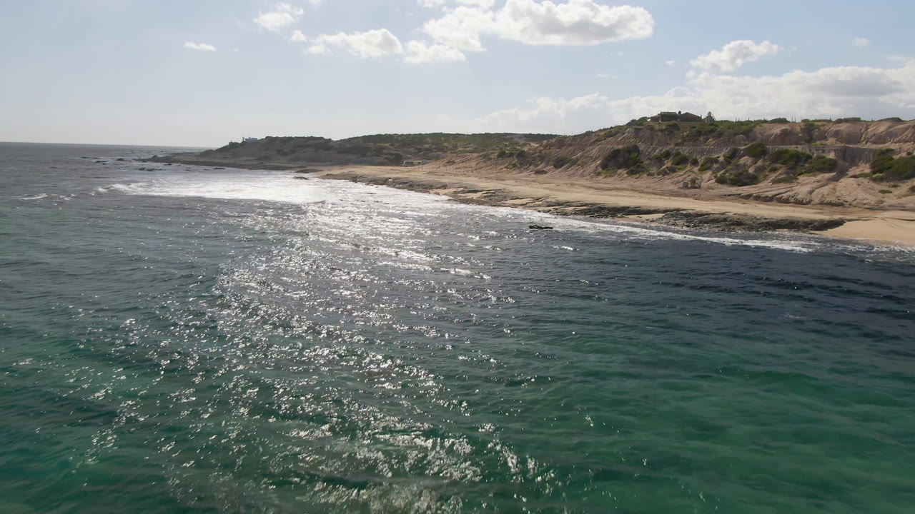 Aerial view over green-blue water toward Shipwrecks Beach in Los Cabos, with bright sun glare on the ocean, surf reaching sand and rocks, and no visible people.