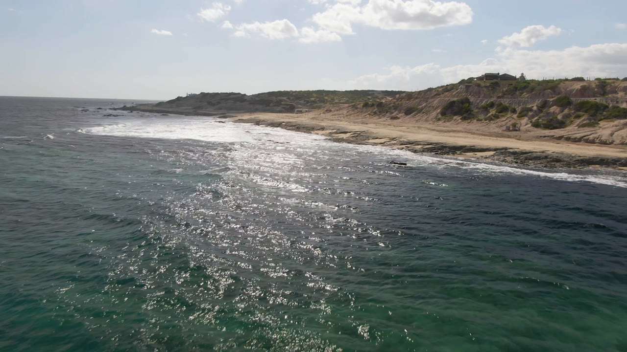 Aerial frame of Shipwrecks Beach in Los Cabos with a broad band of white surf crossing the shore, rocky shallows in the foreground, low bluffs beyond, and no visible people.