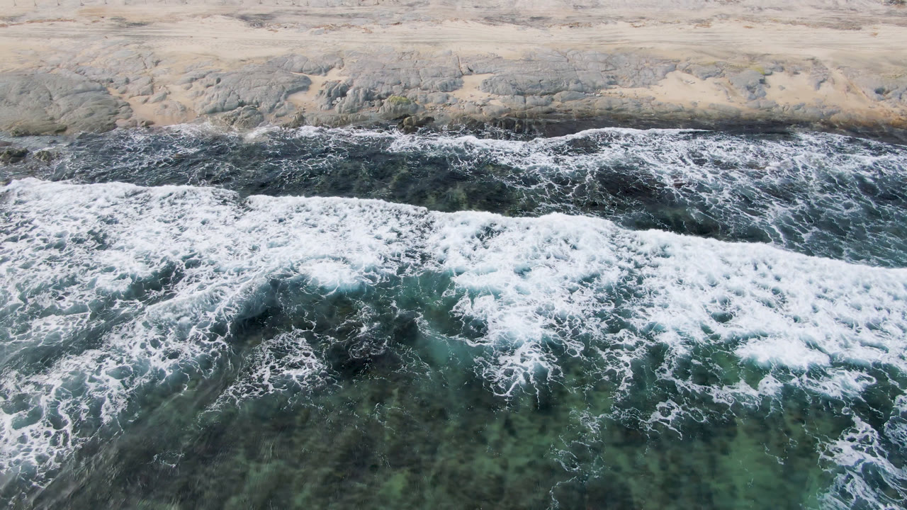 Aerial frame from Shipwrecks Beach in Los Cabos showing white foam spreading over green-blue water below a pale rocky shore, with no visible people.