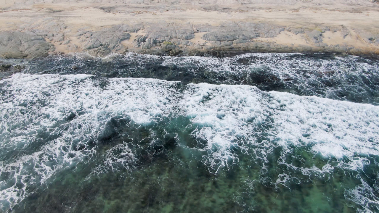 Aerial frame from Shipwrecks Beach in Los Cabos with a broad white wave line crossing dark green water beneath the pale shore, with no visible people.