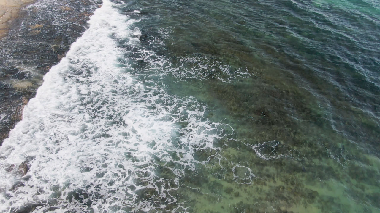 Aerial frame from Shipwrecks Beach in Los Cabos showing a broad band of white foam crossing green-blue water over dark rock, with no visible people.