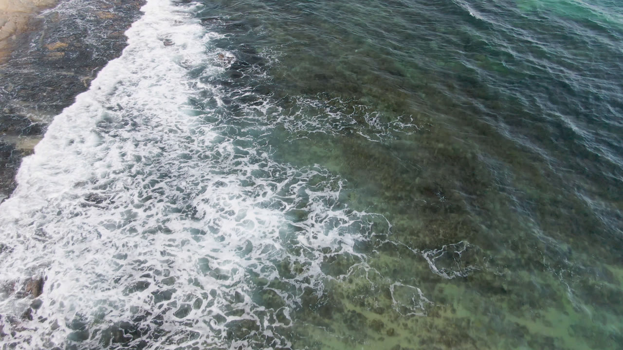 Aerial frame from Shipwrecks Beach in Los Cabos with a curved white wave line pushing through clear green water and darker reef texture, with no visible people.