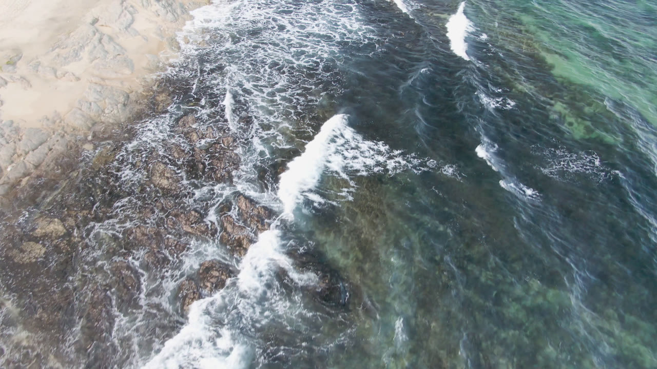 Overhead view at Shipwrecks Beach in Los Cabos shows white surf crossing dark rocks beside pale sand, with green-blue water to the right and no visible people.