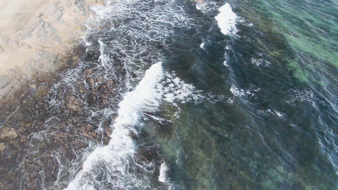 At 0:01, a bright white wave line spreads farther over the rocky outcrop at Shipwrecks Beach, with dark water above and clearer green water along the right edge, no visible people.