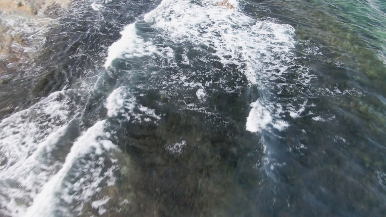 Aerial view at Shipwrecks Beach in Los Cabos showing white surf spreading over dark reef and clear green-blue ocean, with no visible people.