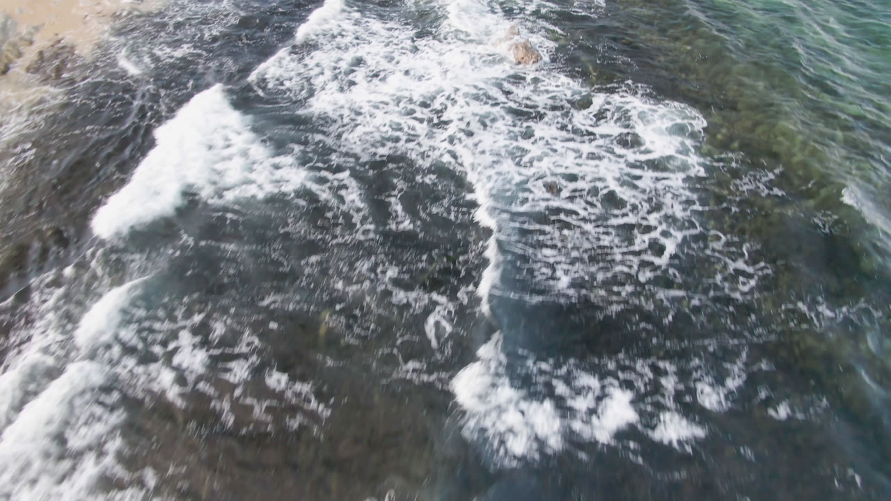 Aerial frame from Shipwrecks Beach in Los Cabos with a broad white foam line crossing dark submerged reef and transparent coastal water, no visible people.