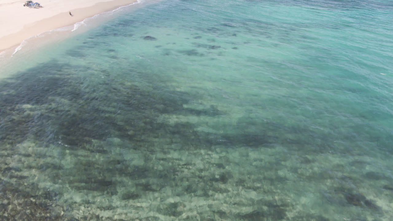 Aerial frame over Shipwrecks Beach in Los Cabos showing turquoise shallows, pale sand, dark underwater patches, a small shipwreck near the upper left, and no visible people.