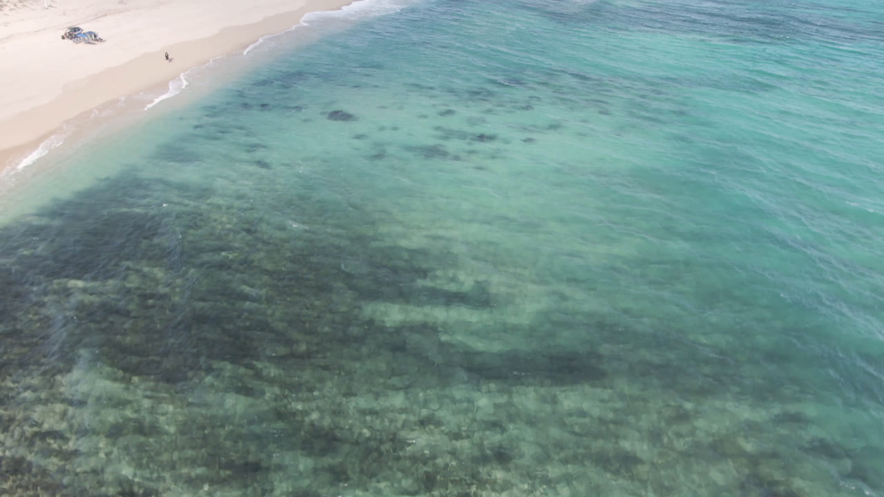 Aerial view at Shipwrecks Beach Los Cabos with a triangular sweep of sand, clear green-blue water, dark reef-like patches below the surface, a small shipwreck on shore, and no visible people.
