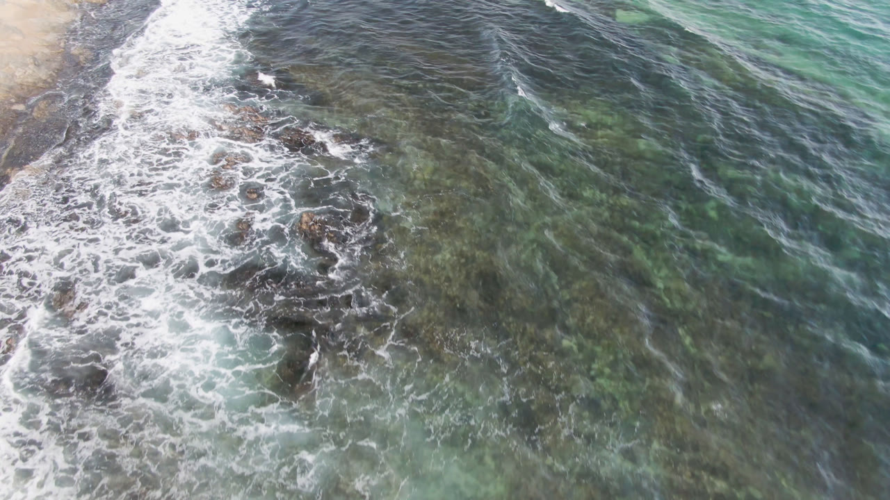 Overhead view at Shipwrecks Beach Los Cabos with white surf washing over dark submerged rocks, green-blue water to the right, pale sand at the upper left, and no visible people.