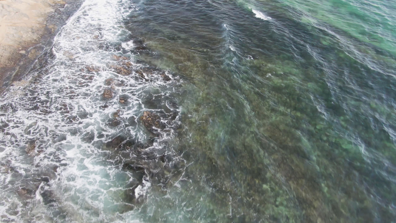 Aerial frame from Shipwrecks Beach Los Cabos showing a wider band of foam crossing rocky shallows, with translucent turquoise-green water filling the right side and no visible people.