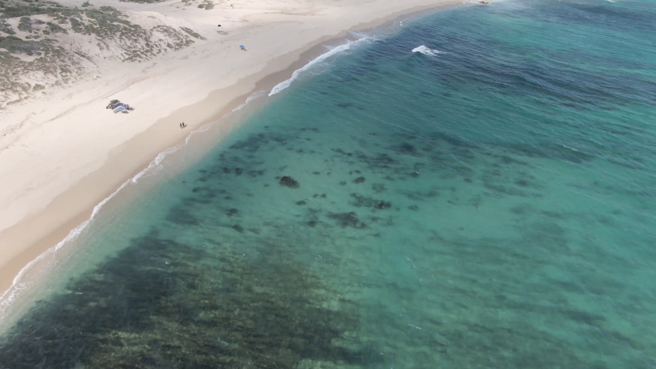 Aerial frame from Shipwrecks Beach in Los Cabos showing turquoise shallows, darker underwater patches, a pale sand shoreline, low dunes, and no visible people at the start of the clip.
