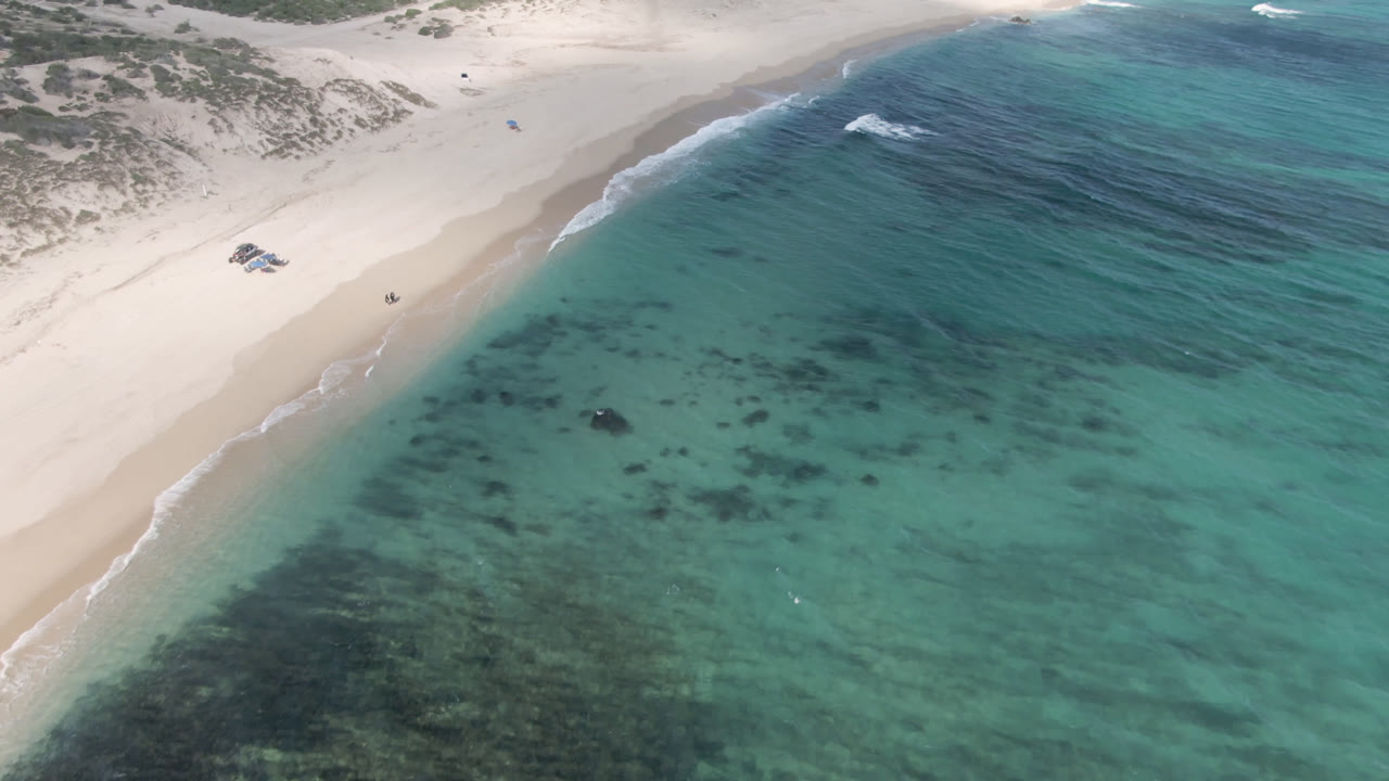 Aerial frame from Shipwrecks Beach in Los Cabos with a curved sand point dividing pale beach from turquoise water, visible dune scrub above the shore, and no visible people.