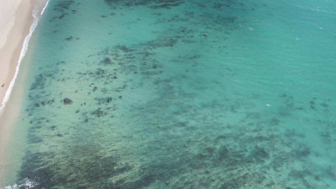 Aerial frame from Shipwrecks Beach showing a narrow strip of sand, foamy water at the shoreline, broad turquoise shallows, scattered dark seabed patches, and no visible people.