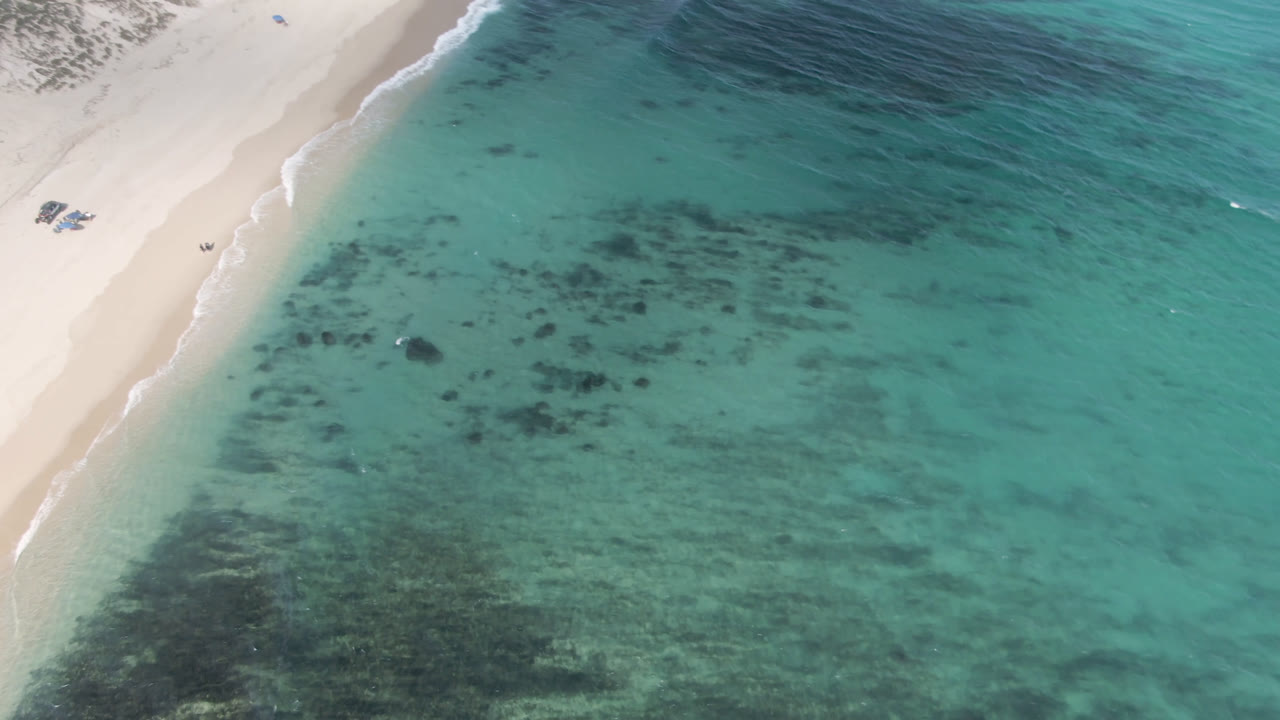 Aerial frame of Shipwrecks Beach in Los Cabos with pale sand at left, turquoise shallows across the center, dark reef patches below the surface, and no visible people.