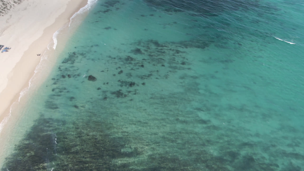 Aerial view over Shipwrecks Beach with the sandy shoreline running diagonally, blue-green water filling most of the frame, scattered dark underwater patches, and no visible people.