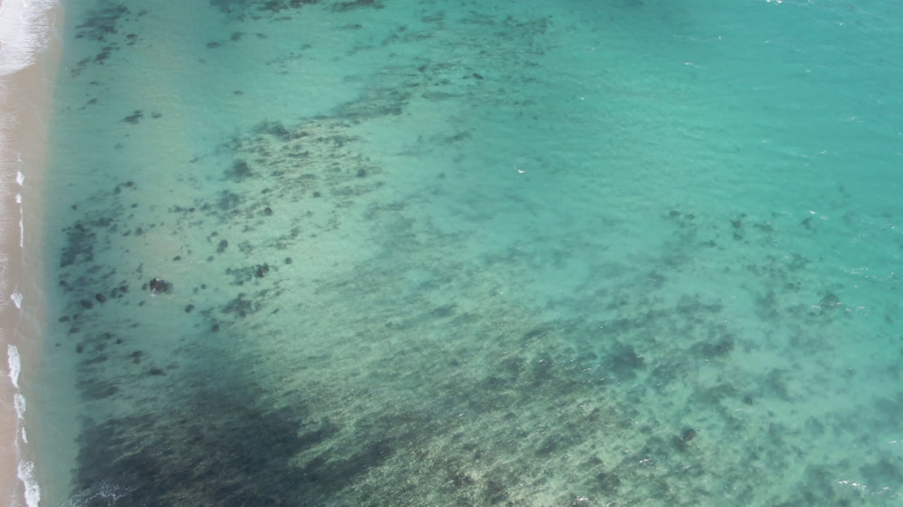 Overhead view at Shipwrecks Beach in Los Cabos with turquoise shallow water, dark underwater patches, pale sand at the left edge, and no visible people.