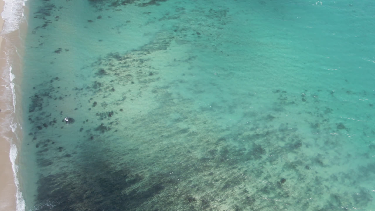 Aerial frame over Shipwrecks Beach showing the vertical sand strip, white wash, turquoise water, dark reef patches, a small dark boat or object near the shore, and no visible people.
