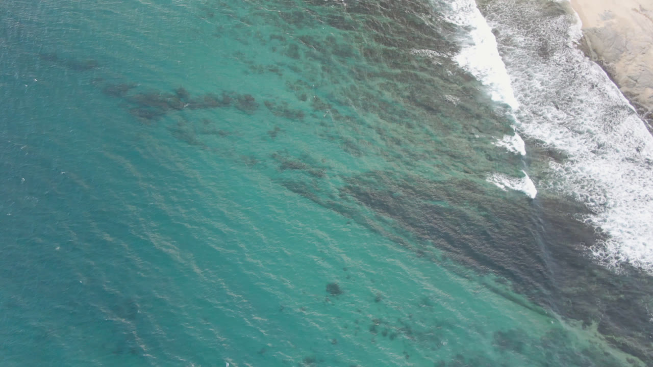 Aerial frame over Shipwrecks Beach in Los Cabos, with blue-green water across most of the image and a narrow band of foamy surf at the right, no visible people.