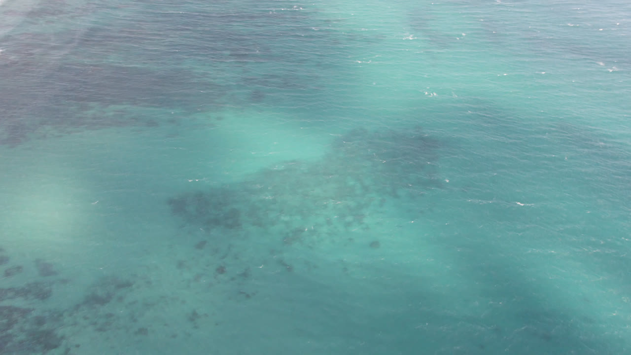 Overhead frame from Shipwrecks Beach in Los Cabos showing teal water, a broad dark patch near the upper left, smaller submerged marks below, and no visible people.