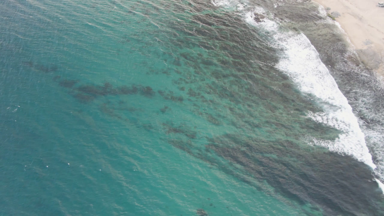 Aerial frame over Shipwrecks Beach in Los Cabos shows teal water crossing dark reef patches, white surf at the rocky sand edge, and no visible people.