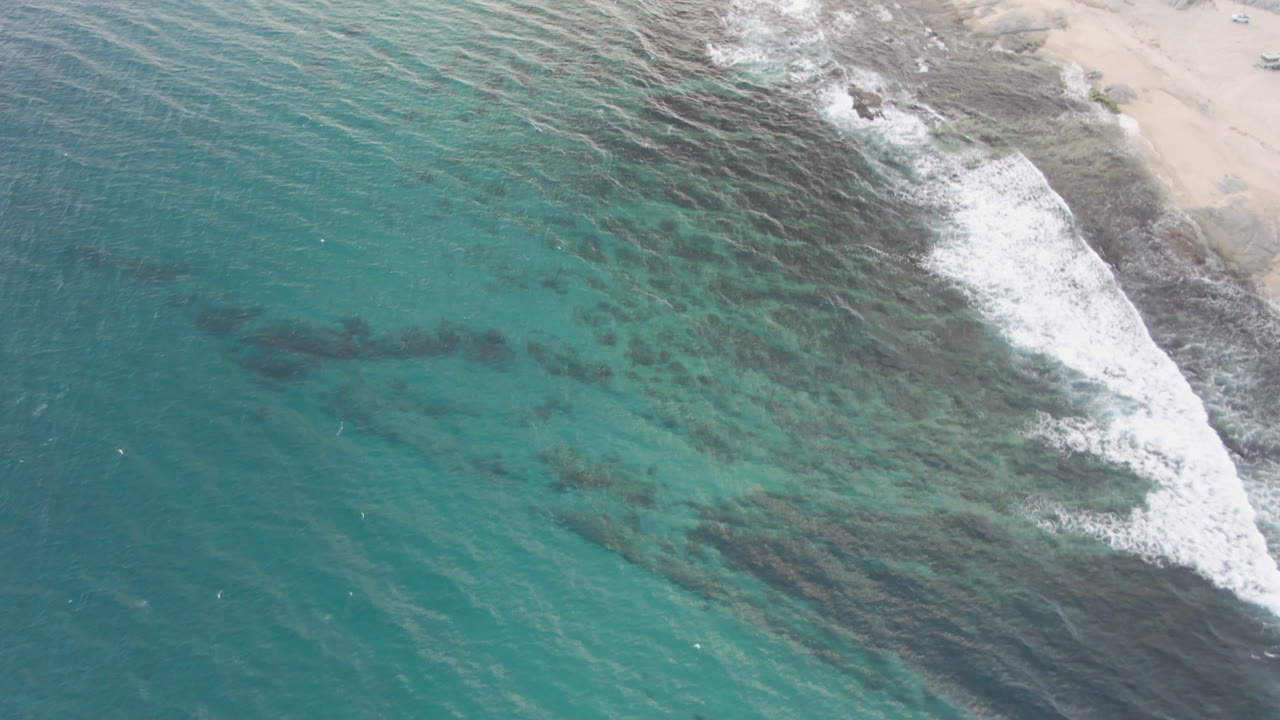 At 0:01, the Shipwrecks Beach aerial shows more of the rocky surf line curving beside pale sand, with turquoise water and no visible people.