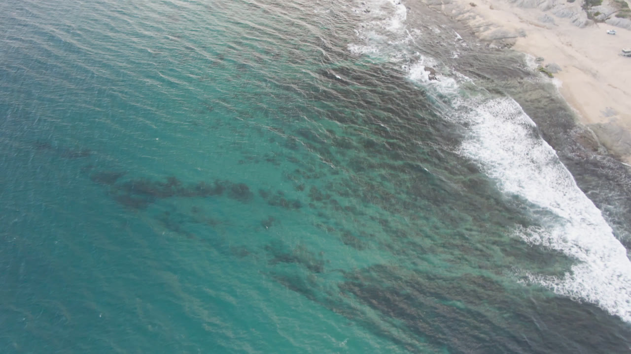 Aerial coastline view with teal water, dark rocks, white foam, and beach edge.