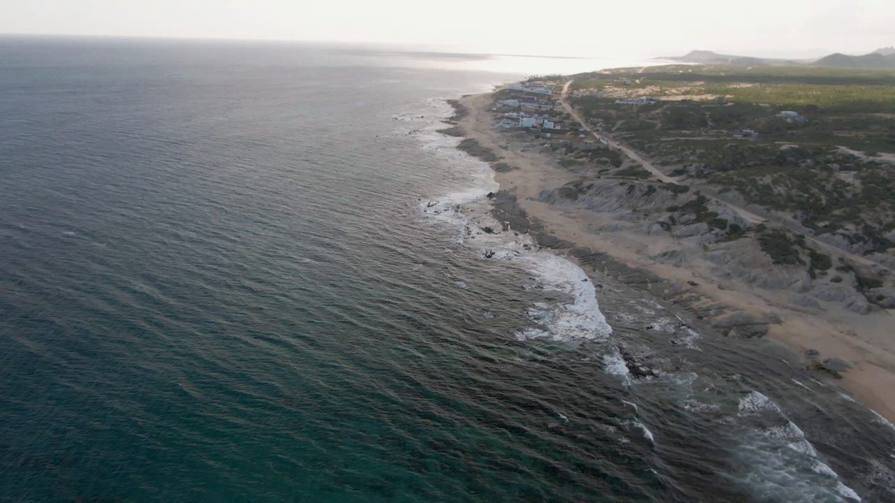 Opening aerial frame over Shipwrecks Beach in Los Cabos, with dark ocean water, foamy surf, sand and rocks, coastal scrub, distant hills, and no visible people.