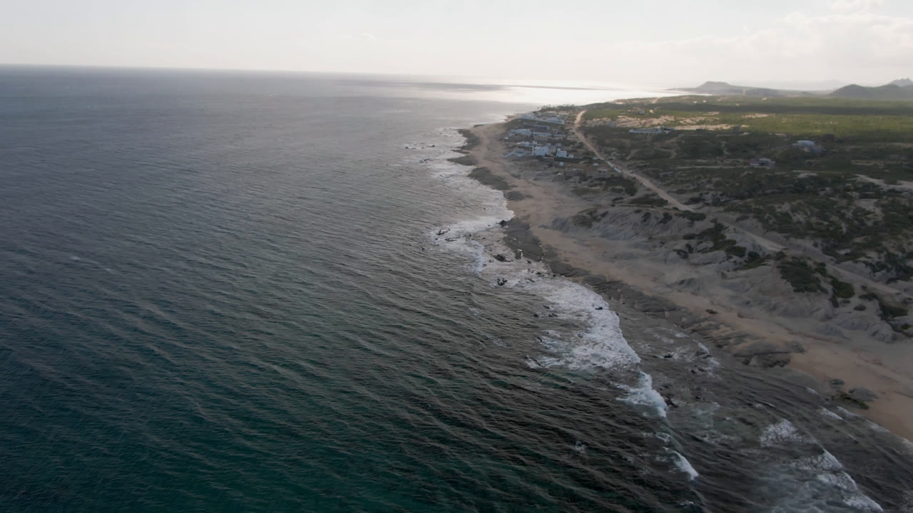 Aerial view along Shipwrecks Beach in Los Cabos, with the surf line slightly farther down the coast, textured water in the foreground, low land beyond, and no visible people.