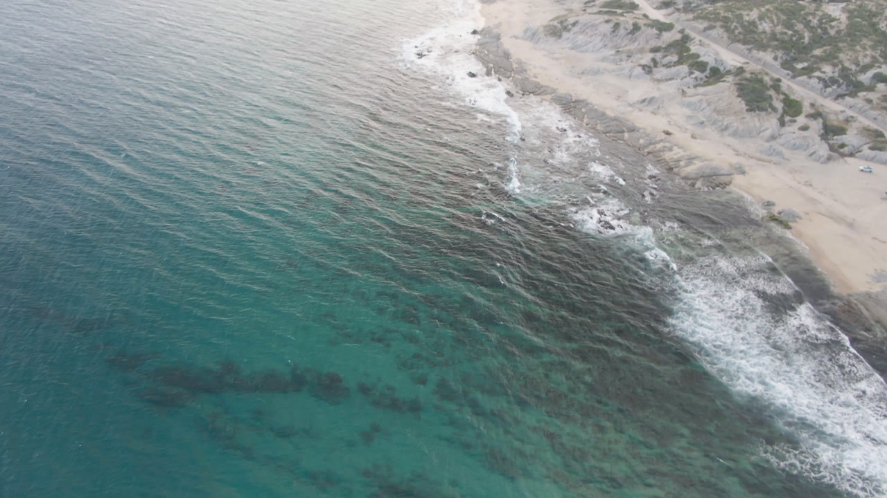 Aerial view over Shipwrecks Beach in Los Cabos, with turquoise water, dark underwater patches, white surf, sandy shore, low vegetation, and no visible people.