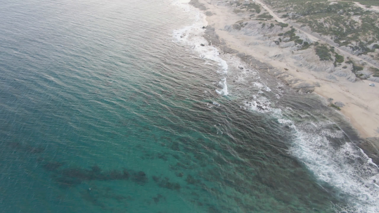 Drone view over Shipwrecks Beach in Los Cabos, showing waves breaking across dark rocks, pale sand at the upper right, open water at the left, and no visible people.