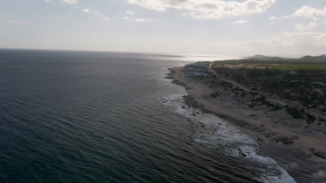 Aerial view over Shipwrecks Beach in Los Cabos, with dark ocean on the left, rocky shoreline curving toward distant hills, scattered clouds, and no visible people.