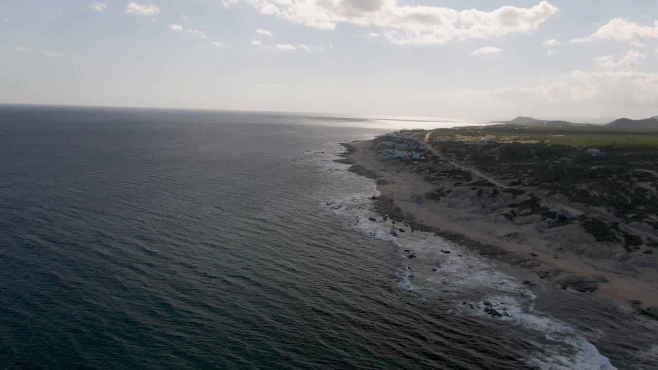 Drone frame from Shipwrecks Beach in Los Cabos looking along a rocky coast, with textured ocean in the foreground, white surf at shore, bright haze near the horizon, and no visible people.