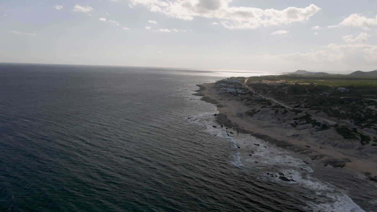 Aerial coastline with ocean, rocks, beach, and low hills under clouds.