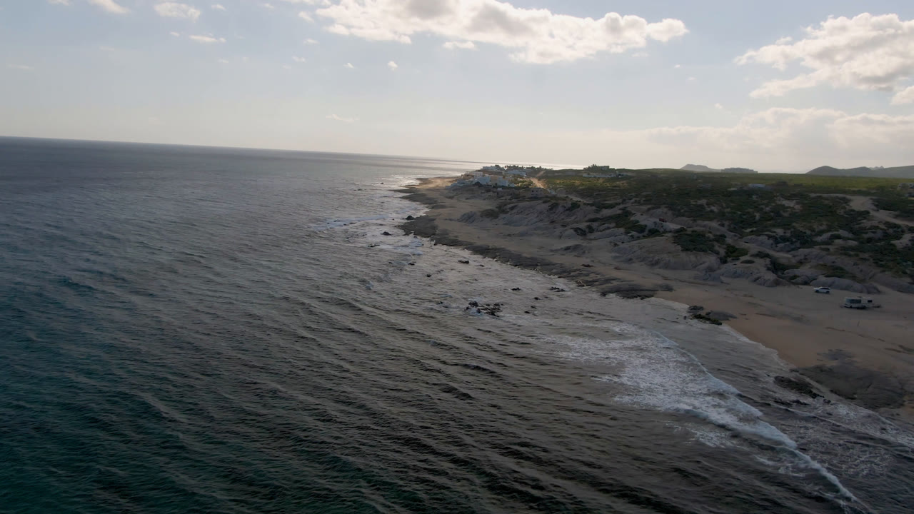 Aerial view over Shipwrecks Beach in Los Cabos, with dark ocean water in the foreground, surf breaking along sand and rocks, low scrubland beyond, and no visible people.